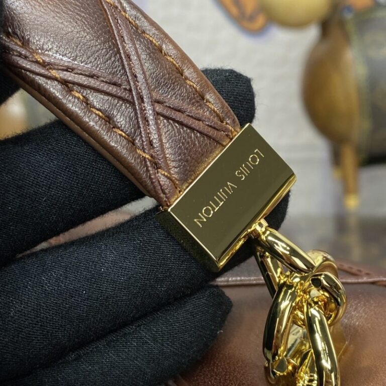 Close-up of a black-gloved hand holding a brown leather strap with gold hardware. The gold clasp is engraved with "LOUIS VUITTON" in capital letters.