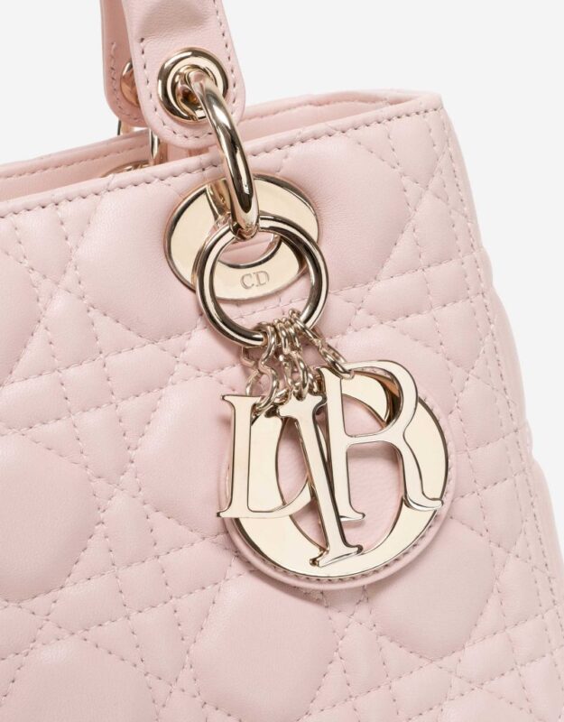 Close-up of a pink quilted handbag with a metal ring and attached gold-tone letter charms hanging from it. The bag features a textured pattern and elegant stitching.