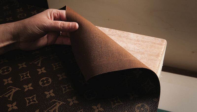 A person holds and lifts the corner of a textured, brown fabric with a distinctive pattern of logos, revealing a wooden surface underneath.