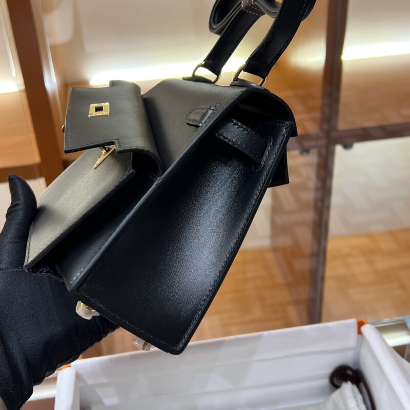 A person wearing a black glove holds the Hermes Kelly Desordre Black handbag at a store. This sleek, black leather bag features a structured design with a small flap pocket on the front. The softly lit background highlights elegant wooden details.