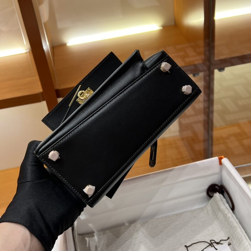 A person wearing black gloves holds a Hermes Kelly Desordre Black handbag, which features a rectangular shape and metallic studs at the bottom. The interior of the bag is visible as it is showcased over an open box, with shelves softly lit in the background.