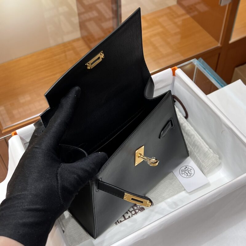 A person wearing black gloves is holding an open Hermes Kelly Desordre Black luxury handbag with gold hardware. The bag is placed on a white fabric inside a box, and the background features wooden display shelves.