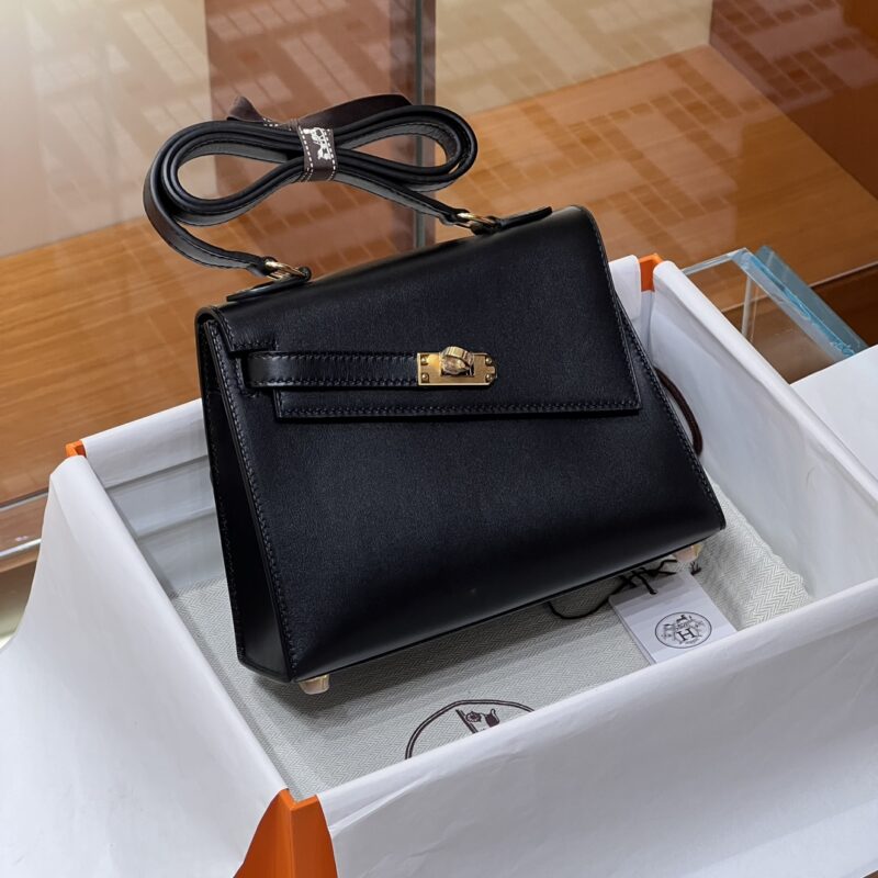 The Hermes Kelly Desordre Black handbag is elegantly presented in a white protective bag inside an orange box. It features a top handle and comes with a detached strap placed neatly beside it. The backdrop highlights a wooden surface.