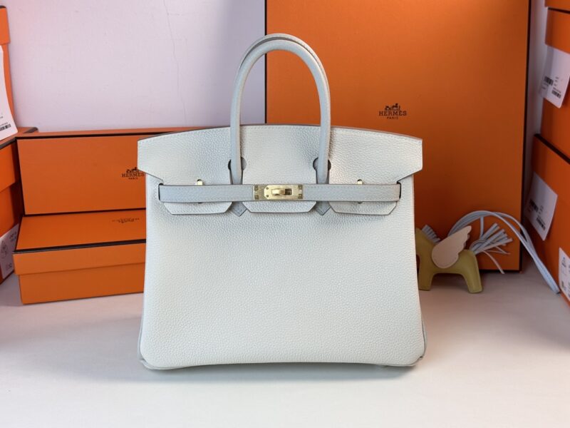 A Hermes Birkin Milkshake White handbag is centered on a table, surrounded by orange boxes featuring brand logos. A small horse-shaped bag charm can be partially seen to the right.