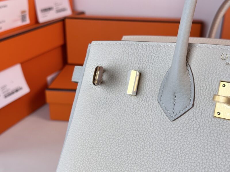 Close-up of the Hermes Birkin Milkshake White handbag, showcasing its white textured design and gold hardware, with a partial view of the handle against a background of stacked orange boxes.