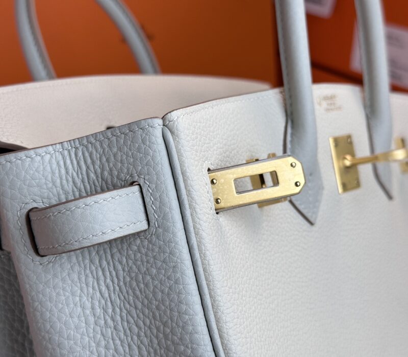 Close-up of the Hermes Birkin Milkshake White luxury handbag, showcasing its light gray leather texture and gold-tone hardware, including a rectangular clasp and handle attachments. The background is set against an orange surface.