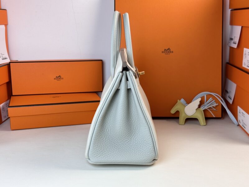 Side view of the Hermes Birkin Milkshake White leather handbag with a textured finish. The background is adorned with several orange boxes and a small horse-shaped ornament, also in orange.