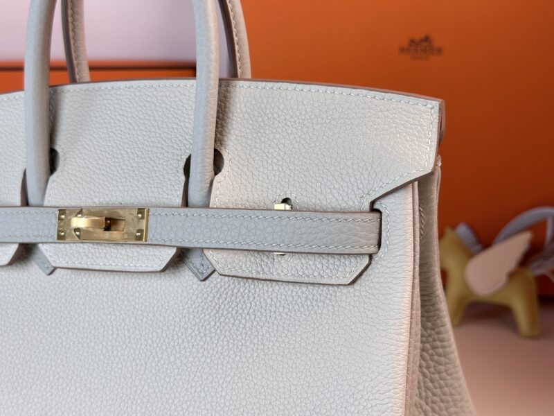 Close-up of the Hermes Birkin Milkshake White leather handbag featuring a gold-toned lock. The bag, with its two top handles, is set against a backdrop that includes an orange box and a small decorative object.