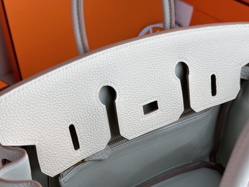 Close-up image of the Hermes Birkin Milkshake White, a luxury leather handbag in a light color, highlighting the top flap with distinct cutouts and stitching details. An orange box is visible in the background.