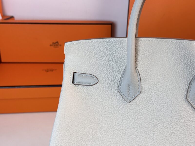 A close-up of the Hermes Birkin Milkshake White handbag with orange boxes in the background. The focus is on the upper corner of the bag, highlighting its stitching and handle design.