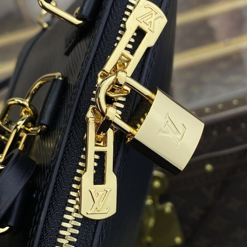 Close-up of a gold padlock attached to a zipper on a luxury black leather bag with gold details. The lock and zipper pulls feature engraved logos.